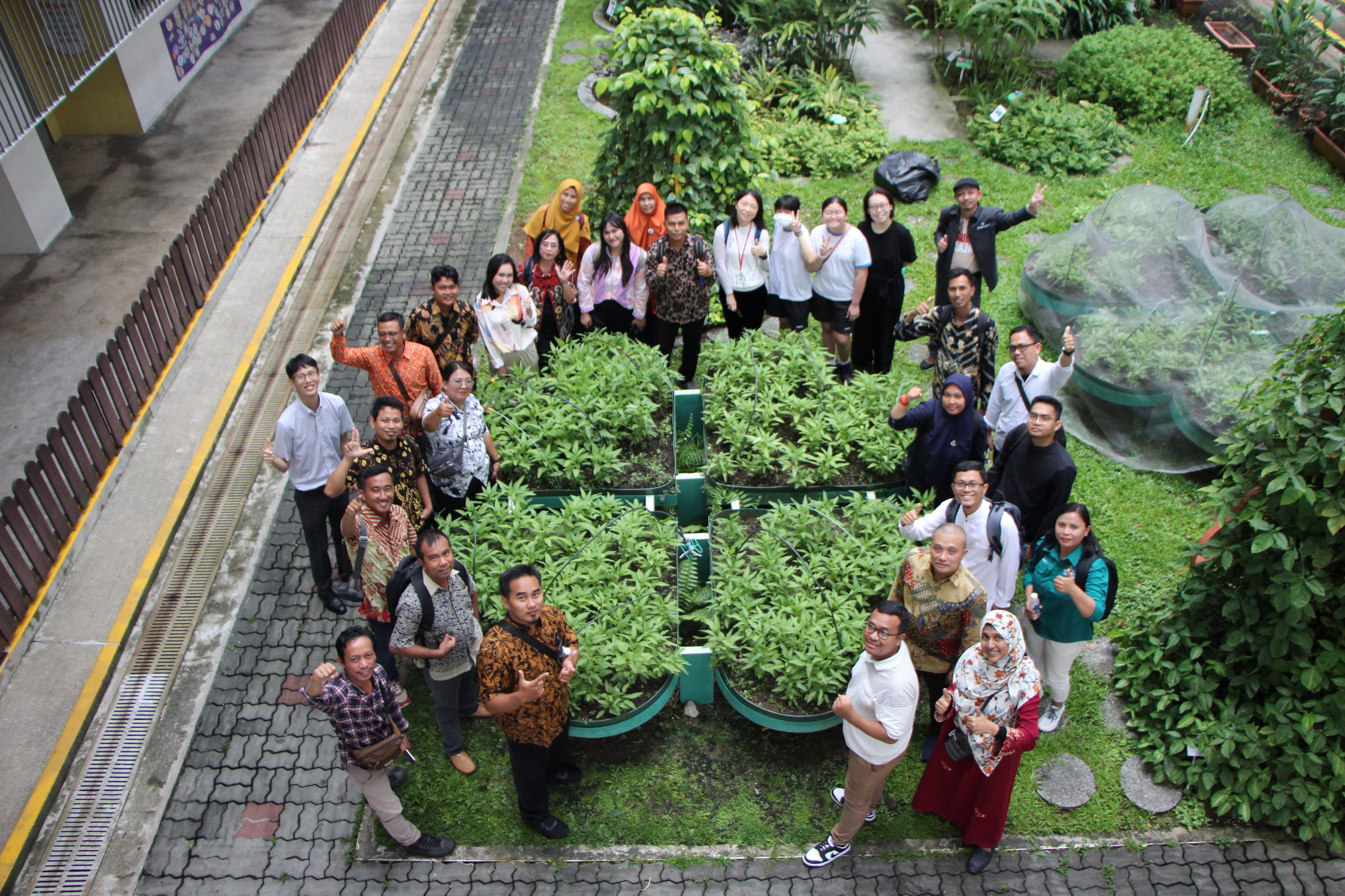 Group of people smiling and waving around four square garden beds in a grassy courtyard, viewed from above.